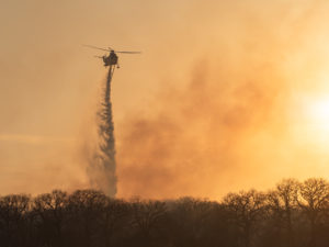 a helicopter is silhouetted against an orange sky and drops fire retardant over Ramsey Fire