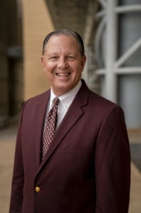 An environmental portrait of a man, Chris Skaggs, in a maroon suit jacket and tie.