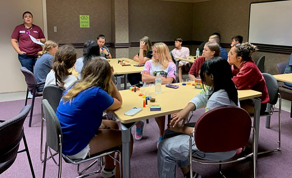 A group of students sit around tables while a woman standing appears to be instructing them.