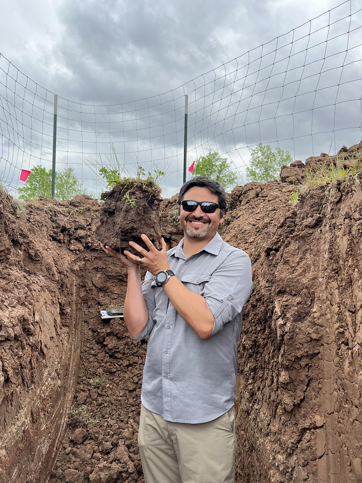 A man in a ditch as deep as he is tall holding a chunk of soil with plants growing in the top of it