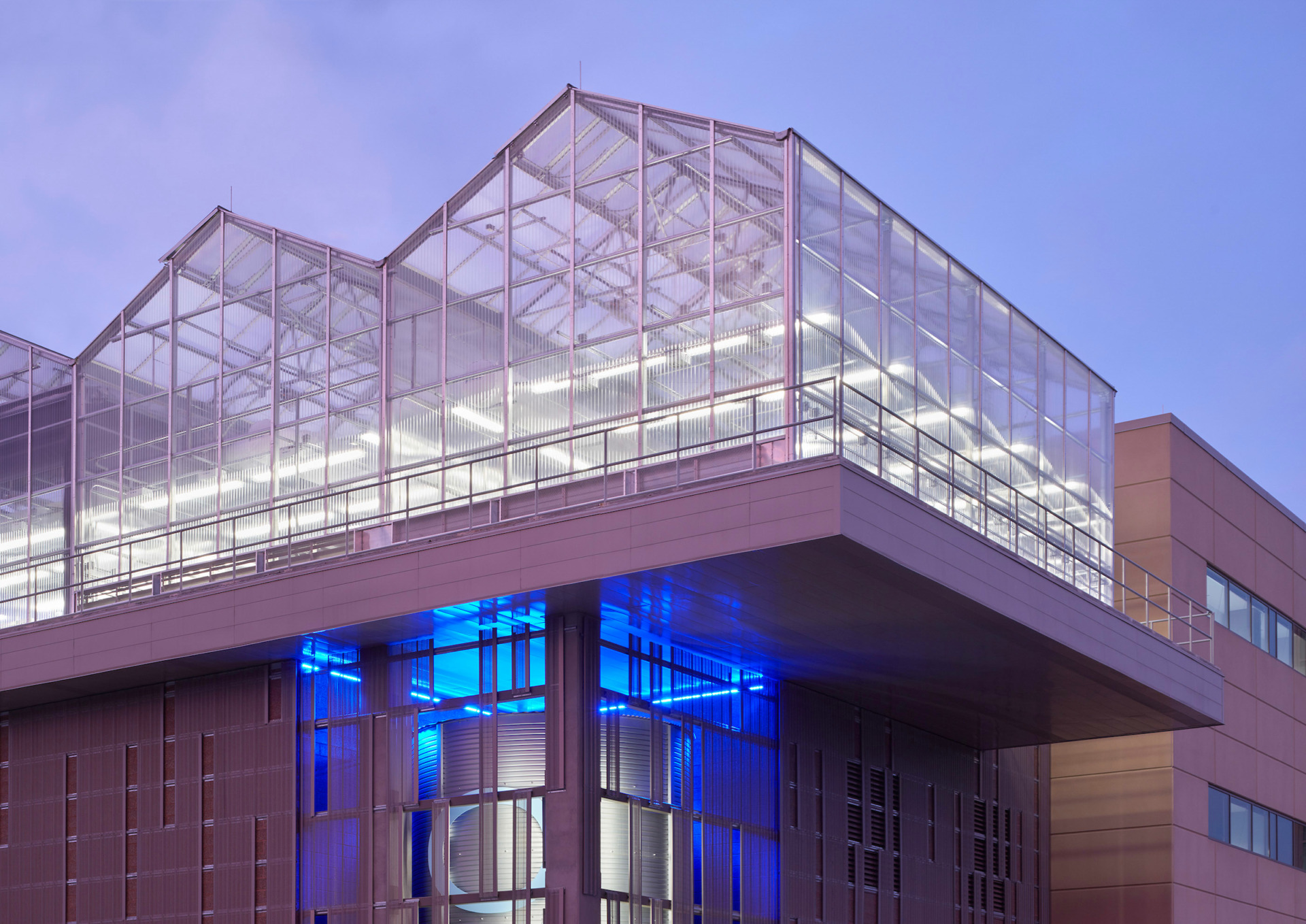 A rooftop greenhouse on a modern building with neon lights emitting from underneath, illuminating a two-story-tall rainwater harvesting cistern.