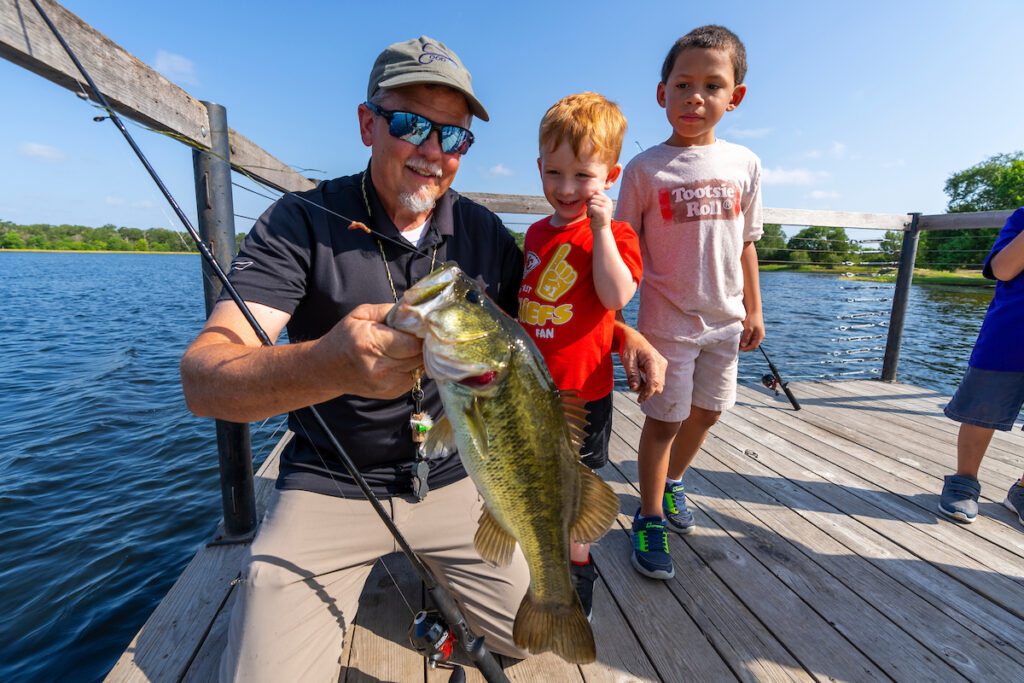 A man holds a large bass after catching it as two little boys look on.