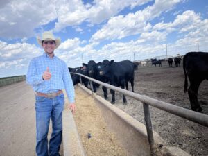 A young man in a cowboy hat, Zach Hoelscher, stands beside a feed bunk in a cattle feedlot with his thumb up during his internship