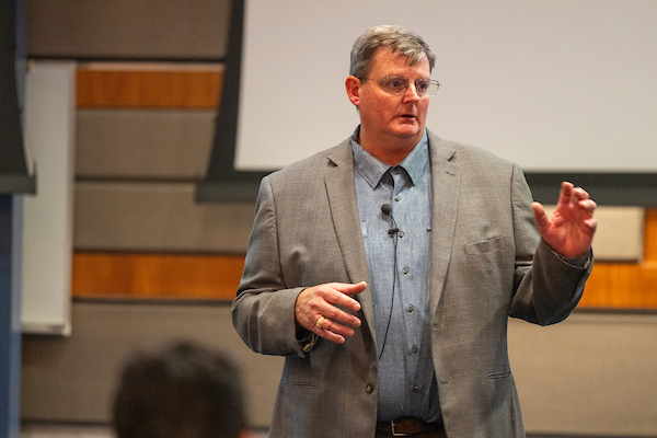 A man in a grey suit and blue shirt confidently presenting to an audience.