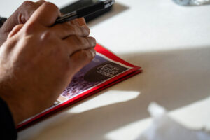A pair of hands holding a pen with a brochure sitting underneath them on a table