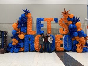 Three women stand smiling in front of a blue and orange baloon arch that reads "Let's Rodeo."