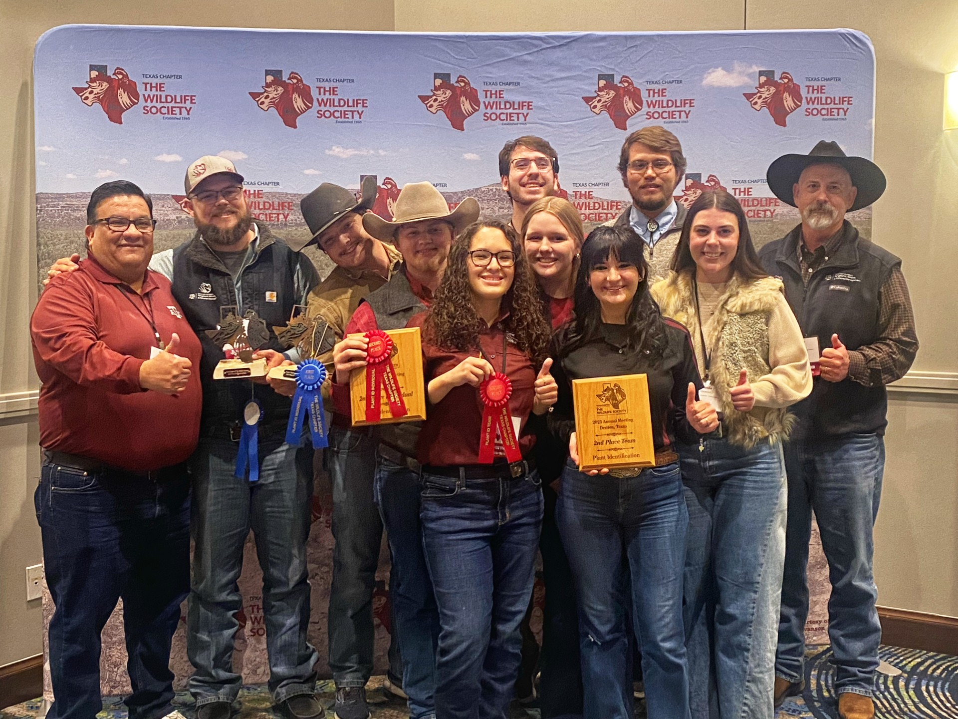 Eleven people stand in front of a banner holding ribbons and award plaques.