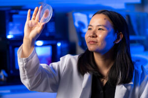 A woman in a white lab coat looks into a Petri dish