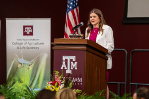 A woman in a white blazer and red dress speaks into a microphone at a podium. The podium has the Texas A&M logo on it, and there is a banner that reads, "College of Agriculture and Life Sciences" beside her.