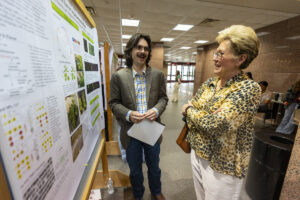 A man in a suit jacket and jeans speaks to a woman in front of a research poster.