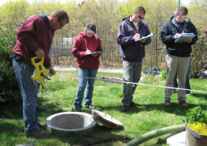 Individuals standing around a septic system cover. One is taking measurements and the others are writing down information