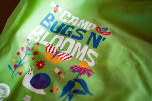 A T-shirt with the words Camp Bugs N' Blooms on it and various insects