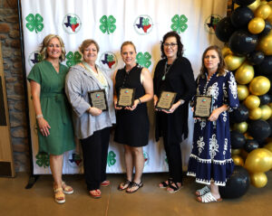 five women stand in from of a 4-H backdrop with an award