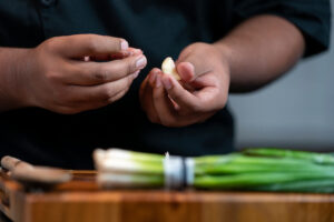 Hands hold a garlic glove while green onions sit on a cutting board.