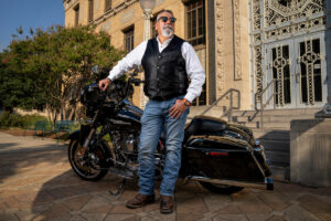 A man wearing a leather vest and jeans stands in front of his Harley motorcycle.