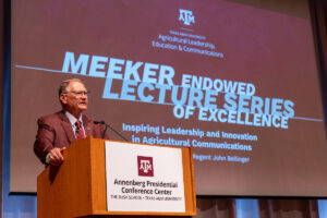 A man in a maroon blazer stands at a podium with a screen behind him that says "Meeker Endowed Lecture Series of Excellence."