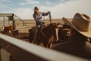 A woman on a horse in a pen with a rope and cattle in the background.