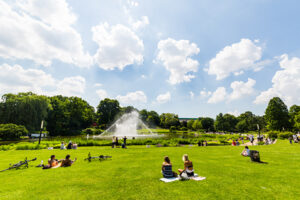 People sit on the grass in a park featuring a pond and a fountain.