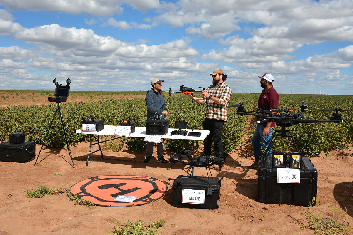 Three men stand in front of a green cotton field with a wide assortment of drones and technical equipment on white tables.