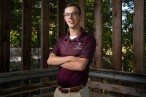 A man wearing a maroon polo stands with his arms crossed in front of wooden planks and a forestry background.