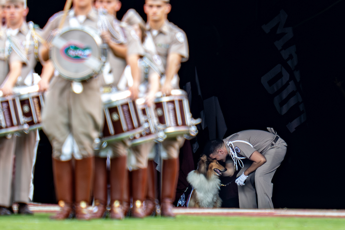 Rows of people in light brown uniforms play musical instruments while a man in the background hugs a dog.