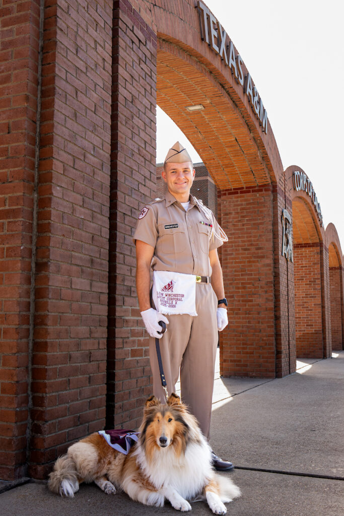 Man in light brown uniform stands with a dog wearing a maroon blanket.