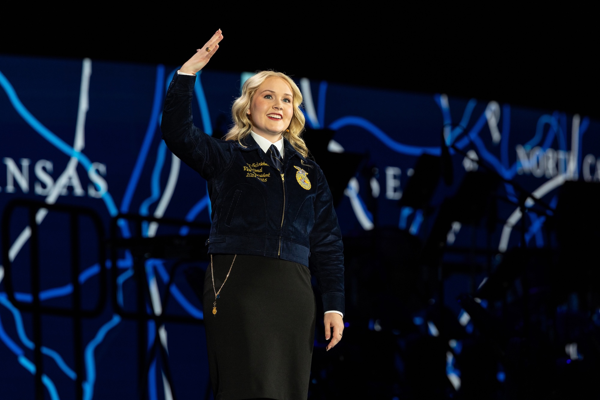 A woman in a blue jacket smiles and waves on a stage.